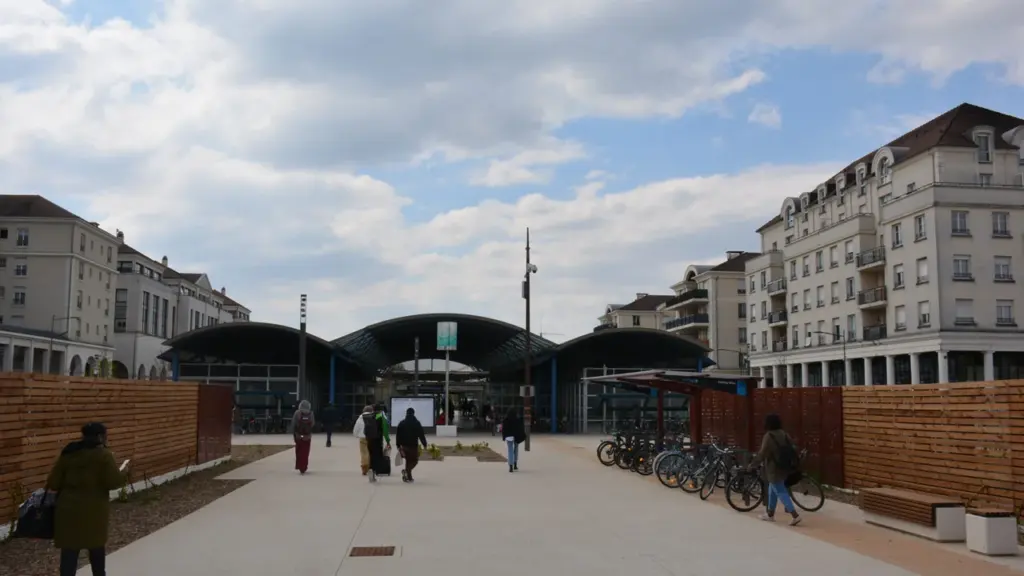 Des piétons marchent sur une esplanade menant à une gare moderne entourée d'immeubles résidentiels.