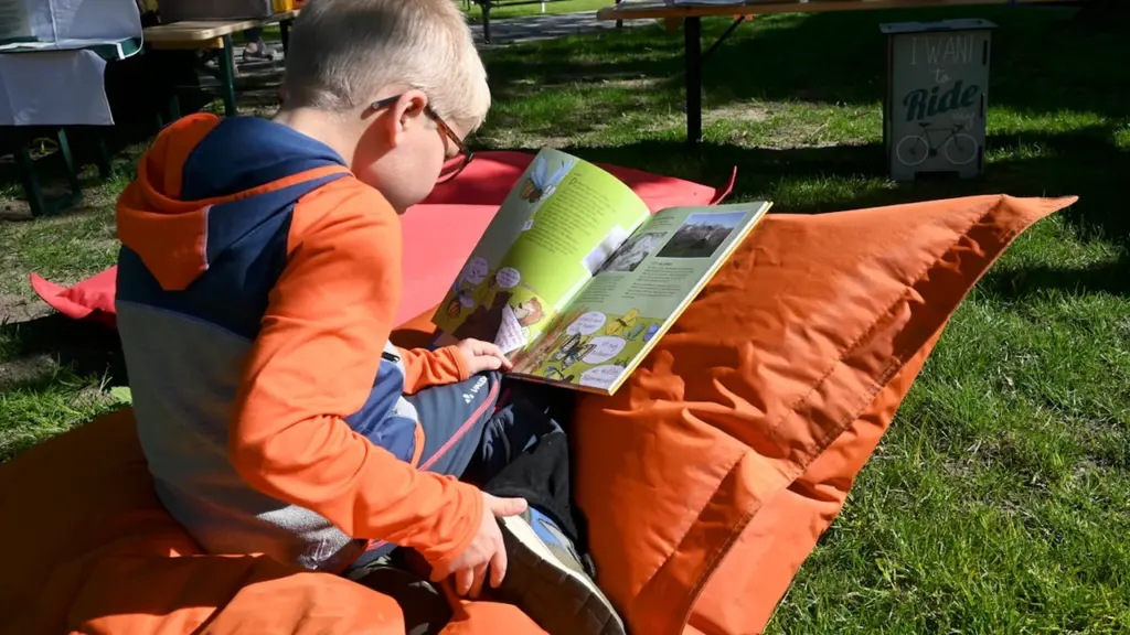 Un jeune garçon lit paisiblement un livre installé sur un coussin orange extérieur.