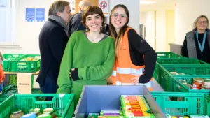 Deux jeunes femmes souriantes posent devant des caisses de nourriture dans une banque alimentaire.