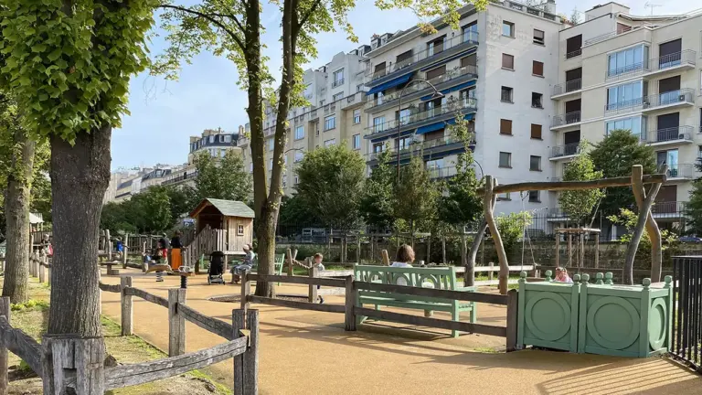 Une aire de jeux urbaine entourée d'arbres avec des équipements en bois et immeubles parisiens.