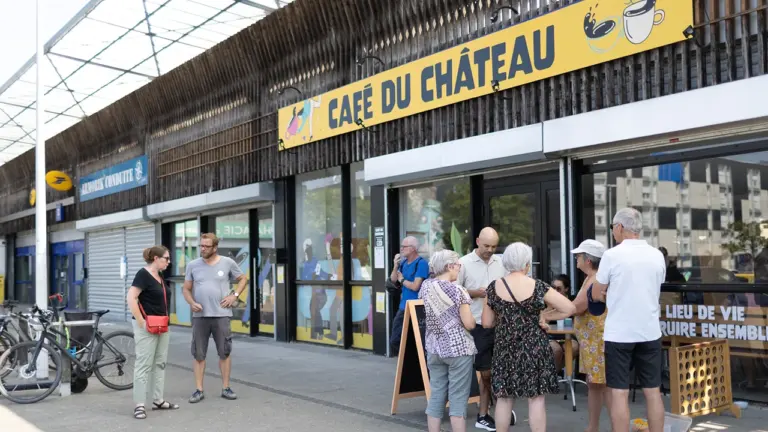 Un groupe de personnes discute devant la devanture jaune du Café du Château extérieur.