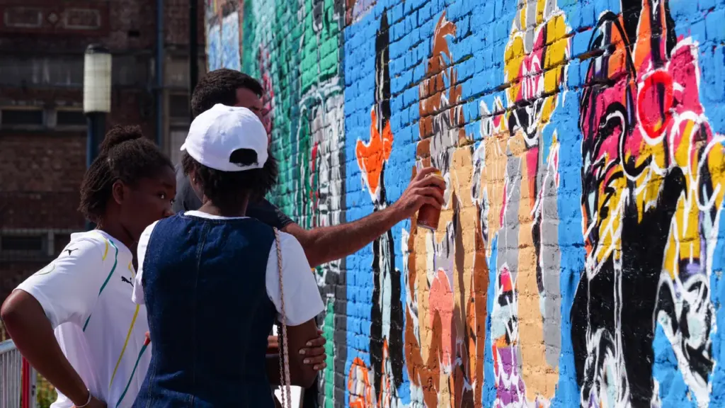 Un artiste graffe un mur en briques coloré sous le regard de deux personnes.