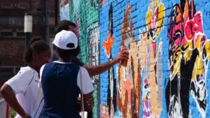 Un artiste graffe un mur en briques coloré sous le regard de deux personnes.