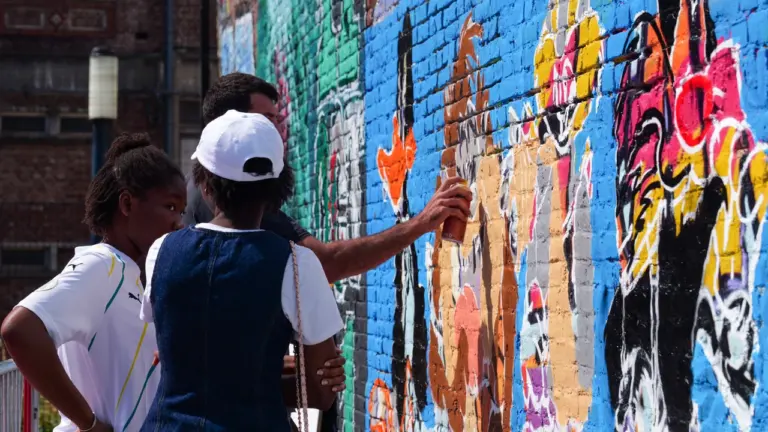 Un artiste graffe un mur en briques coloré sous le regard de deux personnes.