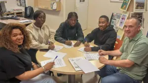Cinq personnes assises autour d'une table ronde signent ensemble des documents administratifs dans un bureau.